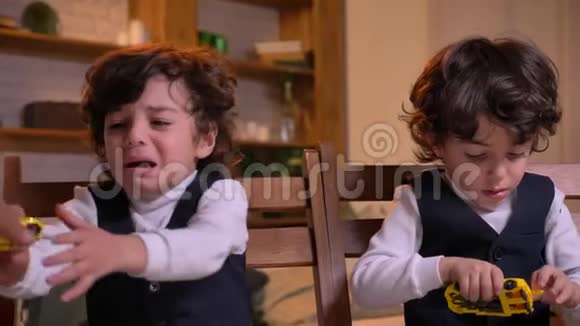两个阿拉伯卷头双胞胎坐在客厅的椅子上哭着要玩具看着镜头的肖像视频的预览图
