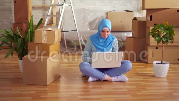 戴着头巾的年轻穆斯林妇女用笔记本电脑坐在盒子旁边的地板上视频的预览图