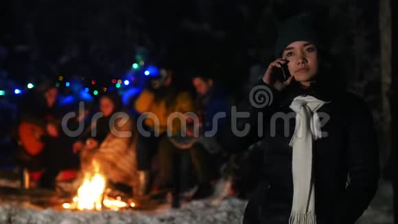 穿着黑色羽绒服的年轻女子站在冬天的森林里在她的电话里交谈一群朋友坐在视频的预览图