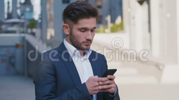 年轻英俊的商人站在大办公楼附近在他的智能手机上输入信息穿着视频的预览图