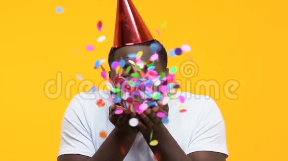 非洲裔美国男性戴着派对帽将纸屑吹进相机生日快乐视频的预览图