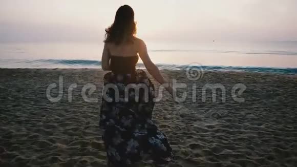 镜头跟随快乐兴奋的女旅行者穿着漂亮的裙子奔向即将到来的海浪在海滩上观看日落视频的预览图
