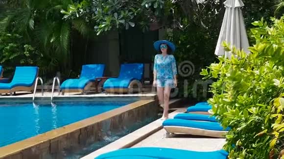 在阳光明媚的日子里穿着蓝色泳衣帽子和太阳镜的漂亮女士带着清澈的水在游泳池里走来走去奢华视频的预览图