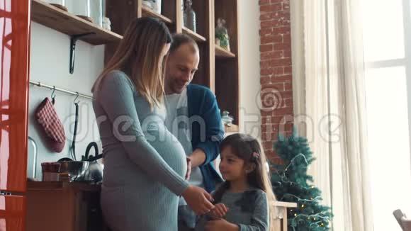 小女孩抱着妈妈怀孕的肚子在厨房里视频的预览图