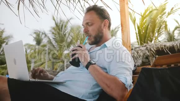 成功的中年男性自由职业者在夏季海滩的躺椅上享受水果饮料同时在笔记本电脑上打字视频的预览图