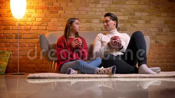 两位年轻的白种人朋友坐在地毯上在舒适的家中喝着美味的热饮料的特写镜头视频的预览图