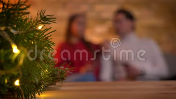 圣诞树明亮的灯光和模糊的年轻夫妇坐在沙发上在背景上交谈视频的预览图