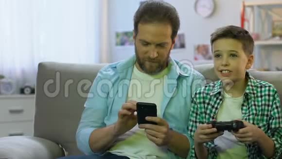带操纵杆玩电子游戏的小男孩父亲在智能手机上滚动视频的预览图