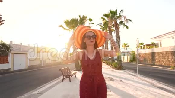 日落时分戴着一顶黄色大帽子的幸福女人沿着棕榈巷散步慢动作视频的预览图