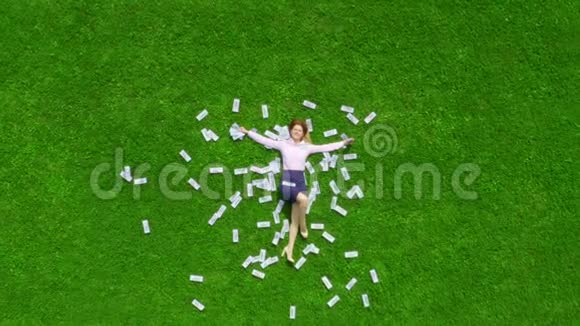 年轻漂亮而成功的女商人躺在一片绿草丛生的草坪上草坪上到处都是钱视频的预览图