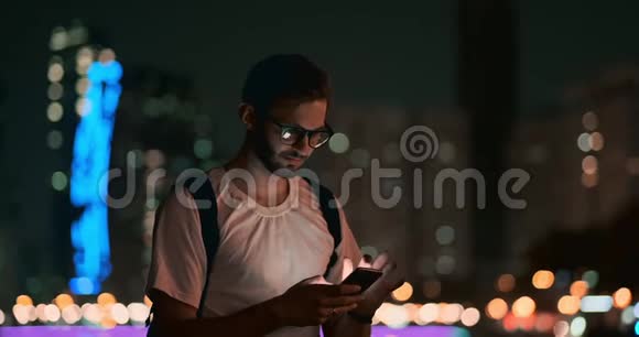 一个戴眼镜的男人在夜城的背景下用智能手机写信息视频的预览图