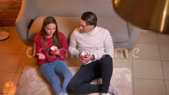 从上往下看快乐的白种人朋友坐在地毯上喝热饮料在舒适的家庭氛围中交流视频的预览图