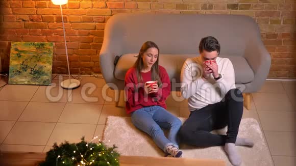 两位年轻朋友坐在地毯上喝着热饮料在圣诞气氛中交流视频的预览图