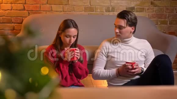 为年轻的白种人朋友在圣诞期间坐在地毯上喝热饮料的特写镜头视频的预览图