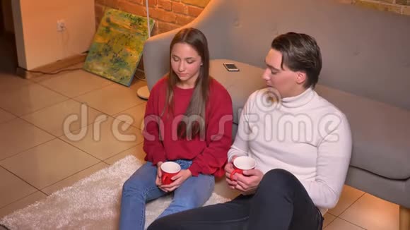两个快乐的白种人朋友坐在地毯上喝着热饮料在舒适的环境中交流他们的特写侧写视频的预览图