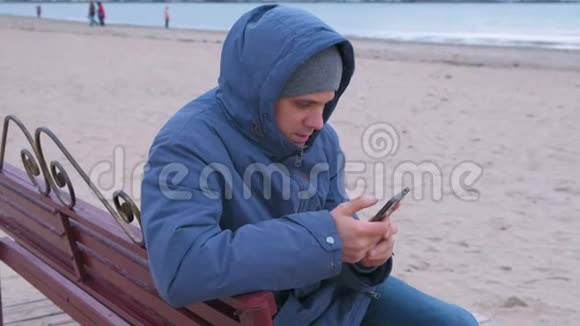 穿着蓝色羽绒服的人坐在沙滩上的长凳上在手机上输入一条信息视频的预览图