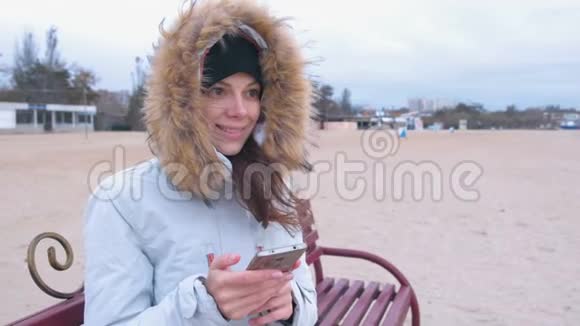 一个穿着白色羽绒服的女人坐在沙滩上的长凳上用手机在社交媒体上打字视频的预览图