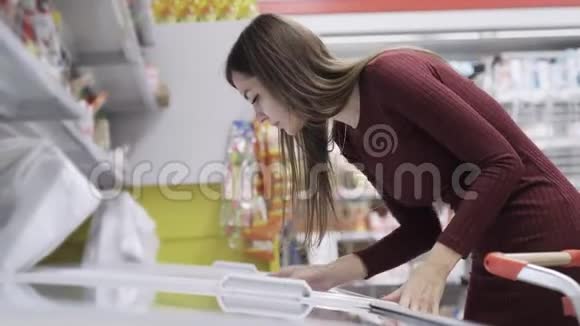 年轻的白种人女人从超市的冰箱里选择冷冻食品视频的预览图
