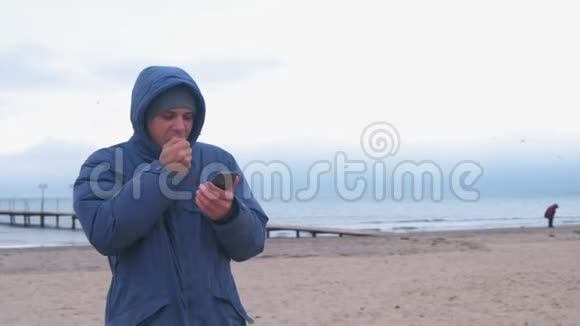 穿着蓝色羽绒服的人在海边的沙滩上用冻僵的手在手机上打字视频的预览图