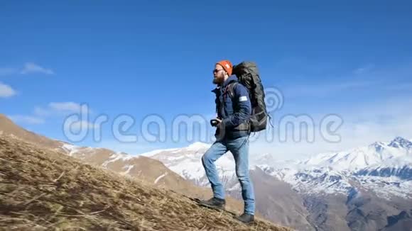 一位留胡子的年轻男性摄影师戴着一顶帽子和太阳镜背着一个大背包脖子上戴着一台照相机爬上了山顶视频的预览图