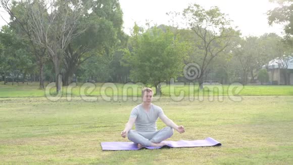 年轻的帅哥在公园里打坐后正在滚动瑜伽垫视频的预览图