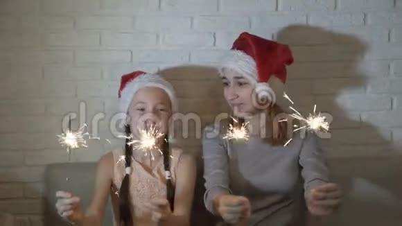 戴着圣诞老人帽子的快乐快乐的孩子们手里拿着点燃的火花挥手坐在沙发上微笑视频的预览图