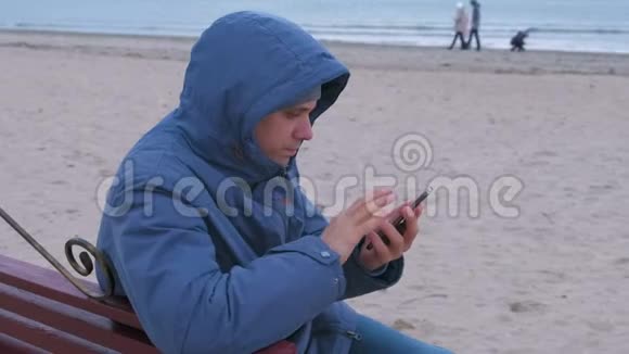 悲伤的男人穿着蓝色羽绒服坐在沙滩上的长凳上打着手机等待答案视频的预览图