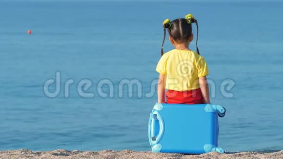 穿着五颜六色衣服的小可爱女孩坐在海边的蓝色手提箱上视频的预览图