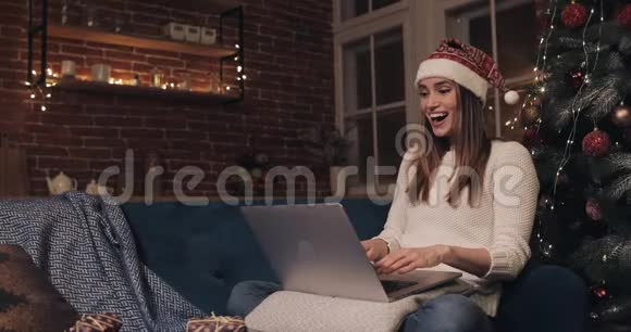 美丽的年轻白种人女孩戴着圣诞帽坐在家背景的圣诞树附近女人用她的笔记本电脑视频的预览图