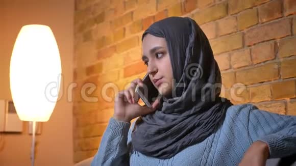 穆斯林年轻女性戴头巾坐在沙发上用电话交谈的特写镜头视频的预览图