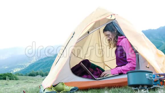 快乐的旅游女性在帐篷里使用笔记本电脑自由职业者在山上的背景下在大自然中打印视频的预览图