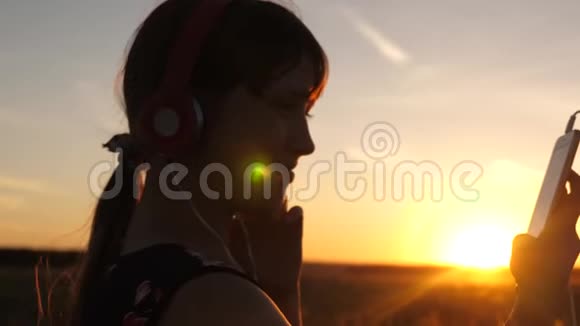 女孩旅行者在智能手机上听音乐在美丽的春天日落的光线下带着耳机的年轻女孩视频的预览图