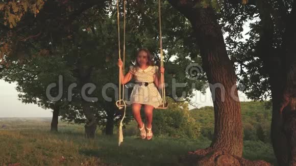 美丽的年轻女孩在阳光下的树下荡秋千和孩子们玩耍孩子们骑着绳子在秋千上视频的预览图