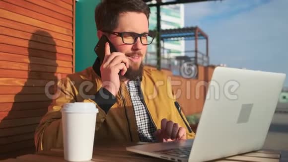 男公民坐在露台上用手机说话在手提电脑上打字视频的预览图