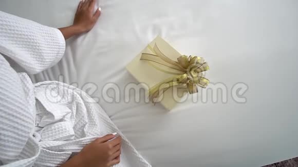 一位穿着白色浴袍的年轻女士坐在卧室的床上她要打开她男朋友送给她的礼物视频的预览图