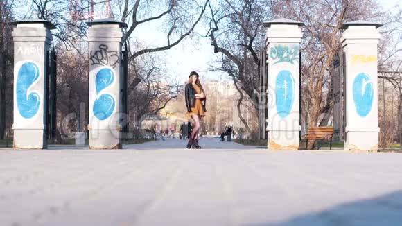 美丽的年轻女子穿着时尚服装戴着黑色帽子脚踝靴和皮夹克站在公园入口附近穿着温暖的衣服视频的预览图