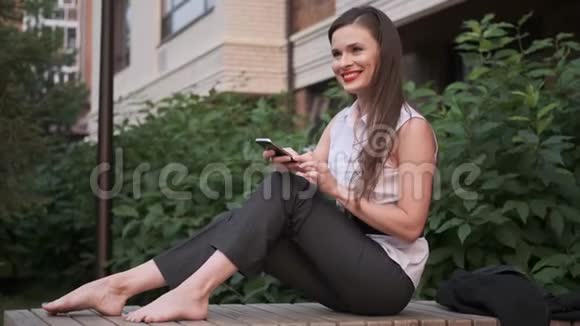 无忧无虑的商务女性赤脚坐在公园里用手机发短信视频的预览图