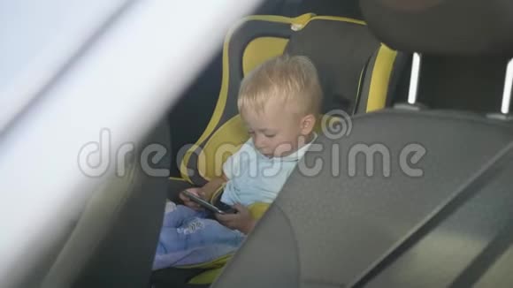 可爱的蹒跚学步的小男孩坐在汽车座椅上从智能手机上观看视频小孩在车里玩智能手机视频的预览图