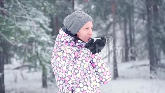 一位冰冻的旅游妇女在下雪的日子里在冬天的森林或公园的背景下从杯子里喝热茶慢慢视频的预览图