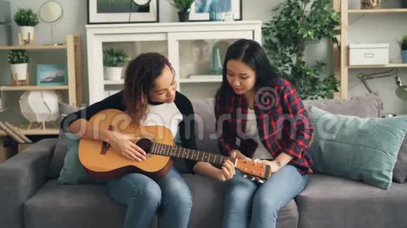 开朗的非裔美国女孩正在家教她的亚洲朋友弹吉他年轻女子坐在沙发上视频的预览图