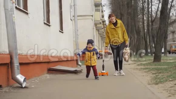 快乐的小男孩带着迷人的微笑沿着路跑去见爸爸妈妈在他身后滚动一辆滑板车慢动作视频的预览图
