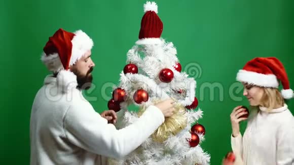 年轻夫妇戴着绿色背景的红帽子装饰圣诞树新年概念视频的预览图