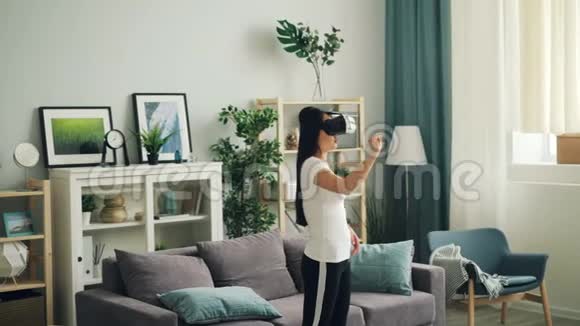 穿休闲服装的年轻女子戴着虚拟现实眼镜双手站在家里现代技术视频的预览图