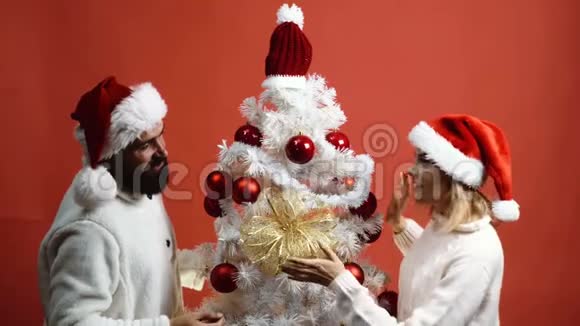 戴着顶帽子的恩爱夫妻在一棵红色背景的圣诞树上有胡子的男人和金发女孩视频的预览图