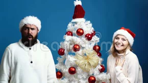 戴着新年帽子长胡子的男人和戴着红帽子的女人站在圣诞树附近表达情感概念视频的预览图