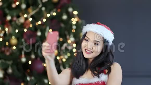 戴圣诞老人套装的亚洲小女孩用智能手机在圣诞树附近自拍视频的预览图