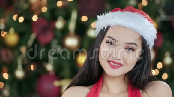 穿着圣诞老人套装戴着圣诞帽穿着圣诞树背景的微笑的亚洲女孩视频的预览图
