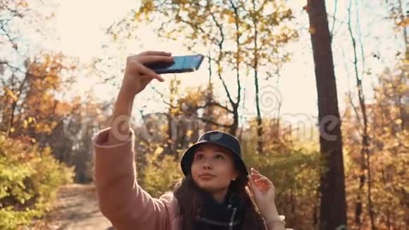 迷人的女孩正在阳光明媚的秋林里用手机自拍微笑着视频的预览图