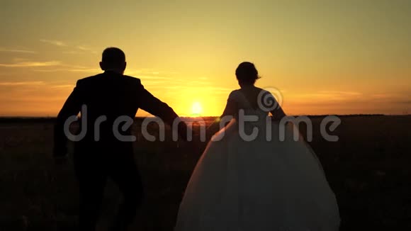 快乐的情侣旅行新娘和新郎在夕阳的照耀下跑过田野恋人们携手在草地上视频的预览图