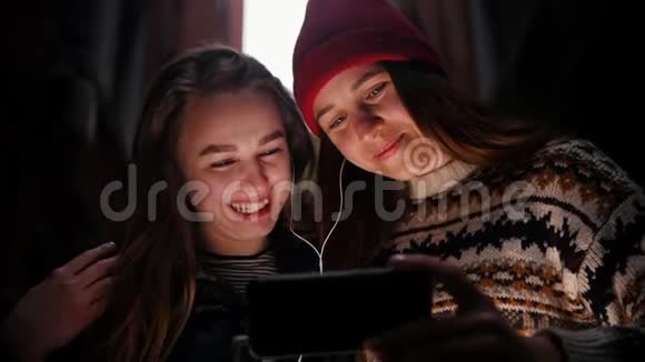两个微笑的年轻女人站在街上用耳机看着手机屏幕视频的预览图